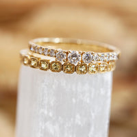 Two gold rings with diamond accents on a white stand against a blurred natural background