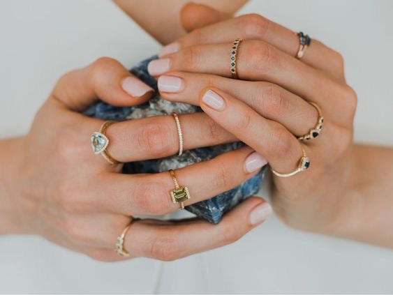 Close-up of hands wearing multiple gold rings with blue and green gemstones on a neutral background