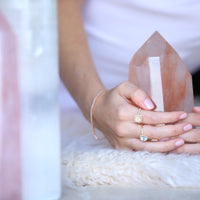 Womans hand holding a quartz tower showcasing 4 rings with Pink gemstones and a gold bracelet. Model is wearing white top and resting on shite sheepskin rug.