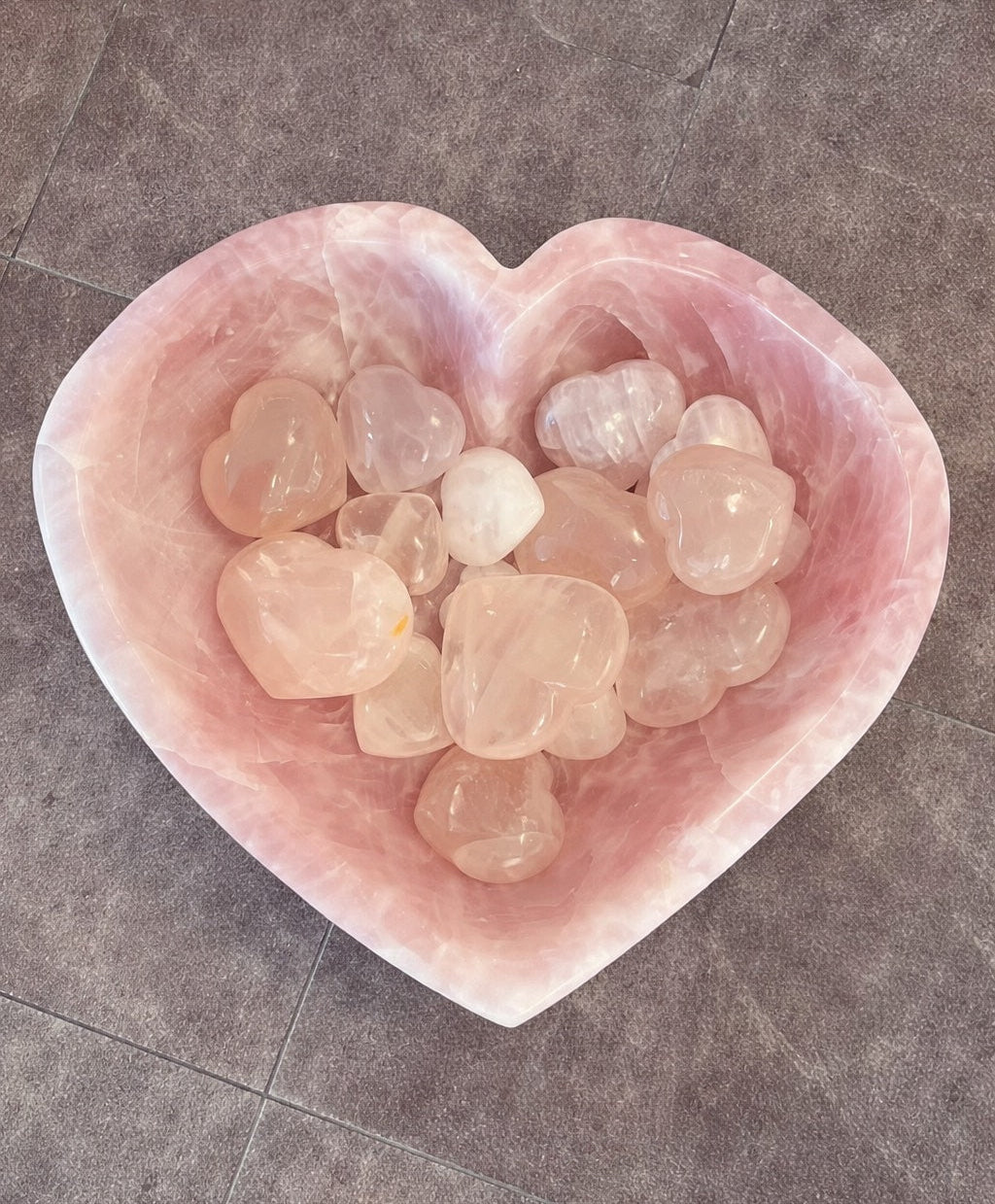 Heart-shaped pink quartz bowl filled with smaller pink quartz hearts on a gray tiled floor