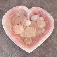 Heart-shaped pink quartz bowl filled with smaller pink quartz hearts on a gray tiled floor